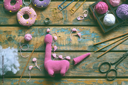Amigurumi dinosaur toy made of pink yarn with legs and tail visible surrounded by wool balls and tools on colorful wooden background in crafting scene, step 3 - to sew all details of toy