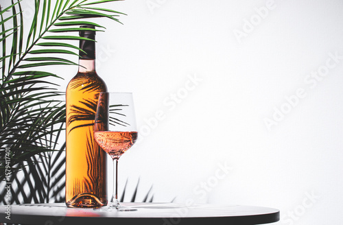 Fototapeta Naklejka Na Ścianę i Meble -  Rose pink wine served in an elegant glass beside a bottle against a light background with green palm leaves