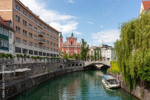 A picturesque view of the Ljubljanica River flowing through the charming city center of Ljubljana, Slovenia, under a bright, partly cloudy sky