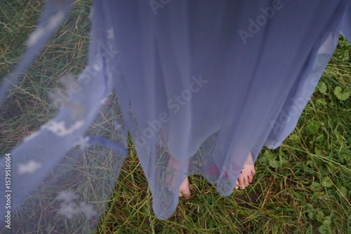 Mädchen mit blau-grauem langen Kleid steht barfuß auf grüner Wiese in der Natur am Abend im Sommer