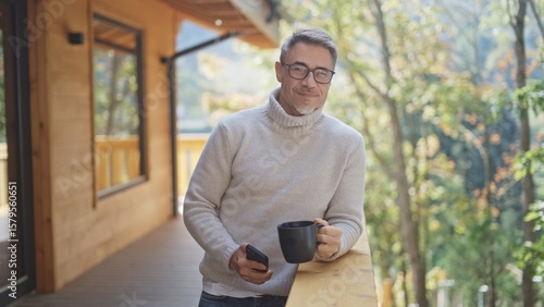 Relaxed man drinking coffee using mobile phone on balcony of modern wooden house in mountains. Portrait of happy mature businessman enjoying nature and peaceful moment of vacation, smiling.