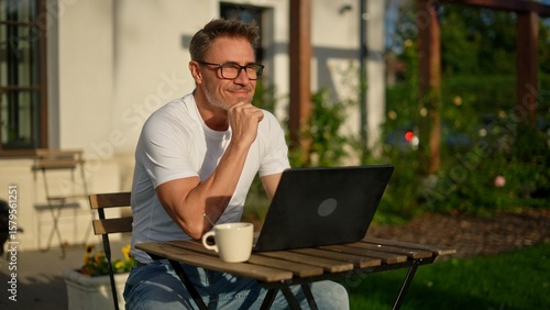 Happy businessman working remotely in garden. Mature man using laptop computer outdoor in morning with coffee. Male entrepreneur freelancer tele working online in home office. Digital business life.