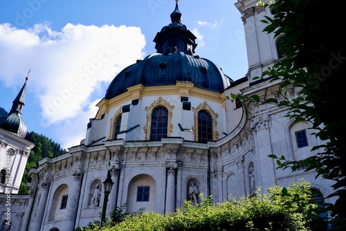 Barocke Klosterkirche mit Kuppel vor blauem Himmel in Alpenkulisse