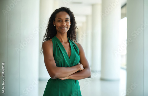 Free Download Empowered African American woman wearing green dress stands confidently in modern corporate building lobby. Arms crossed, smiling, exuding professionalism, intelligence. Represents success, ambition, - GFXLoad