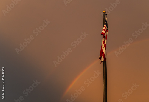 American flad stands proudly with rainbow in background as darkness approaches at sunset. 