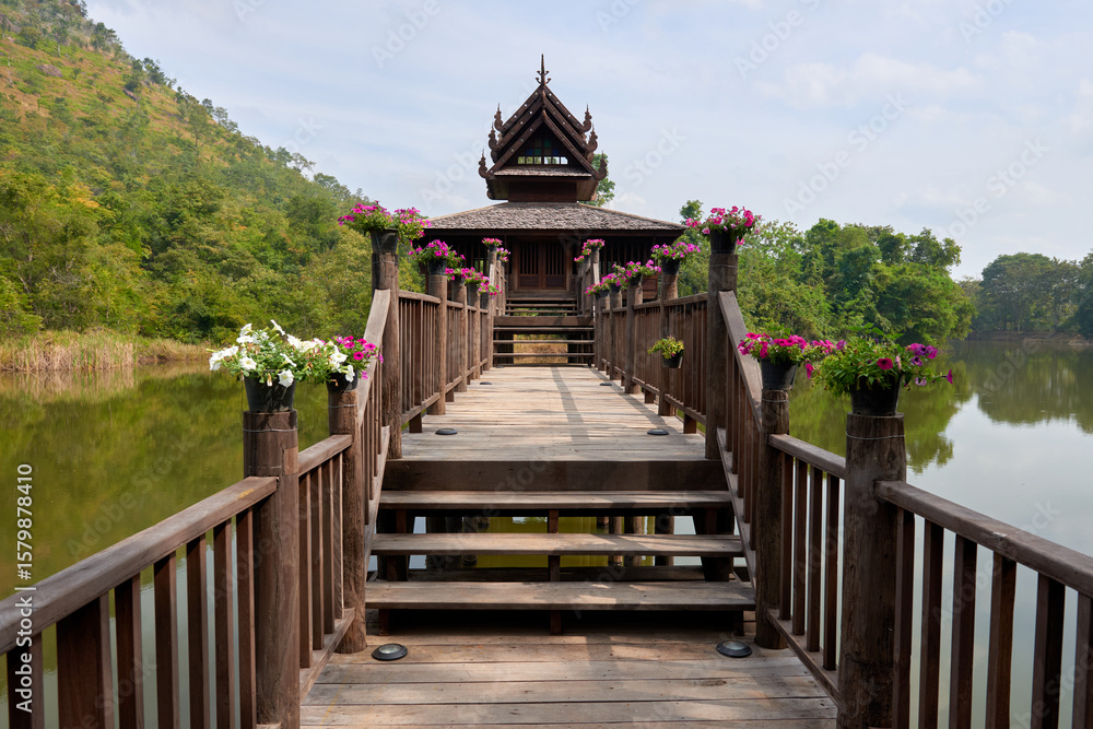 Obraz premium Stunning Wooden Bridge Leading to a Traditional Structure by a Serene Lake Surrounded by Lush Greenery