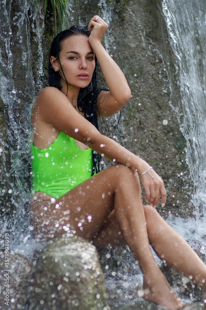 Obraz premium Attractive woman with perfect body posing near waterfall. Wet sexy woman. Portrait of a girl in bikini resting on a paradise beach. Beautiful young woman in water. Wet sexy slim body.