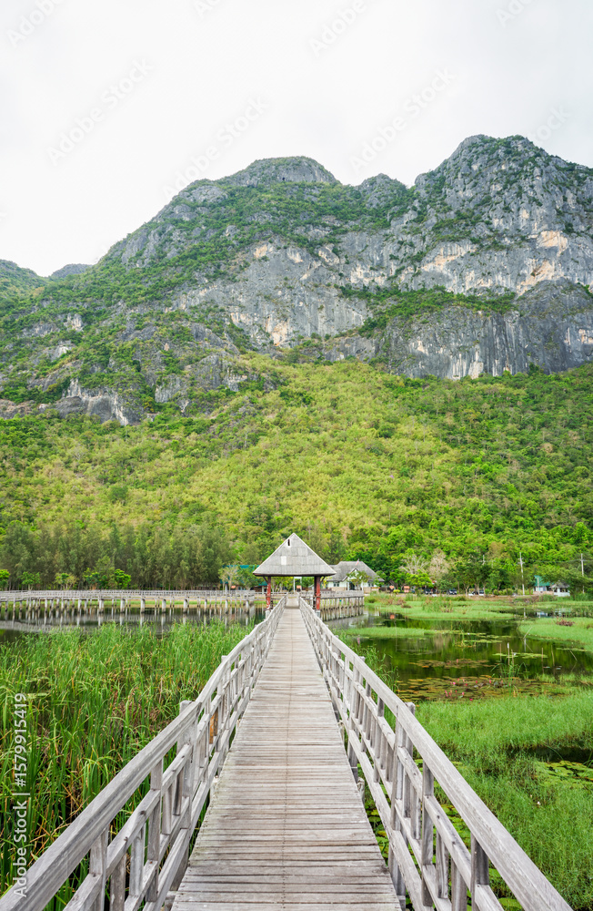 Fototapeta premium Wooden Boardwalk and Pavilion Amidst Bueng Bua Wetlands. Wood walkway extends into the serene wetlands at Sam Roi Yot, surrounded by tall reeds and lily pads, distant pavilion and mountain