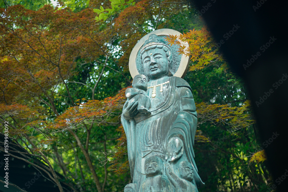 Fototapeta premium A peaceful bronze statue of Kannon, the Buddhist goddess of mercy, holding a child at Myoen-ji Temple in Japan. Surrounded by colorful trees in a tranquil garden setting, symbolizing compassion and sp