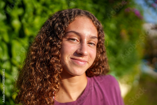 Hispanic teenage girl with curly hair standing confidently in sunlit park, expressing vibrant youth and genuine happiness