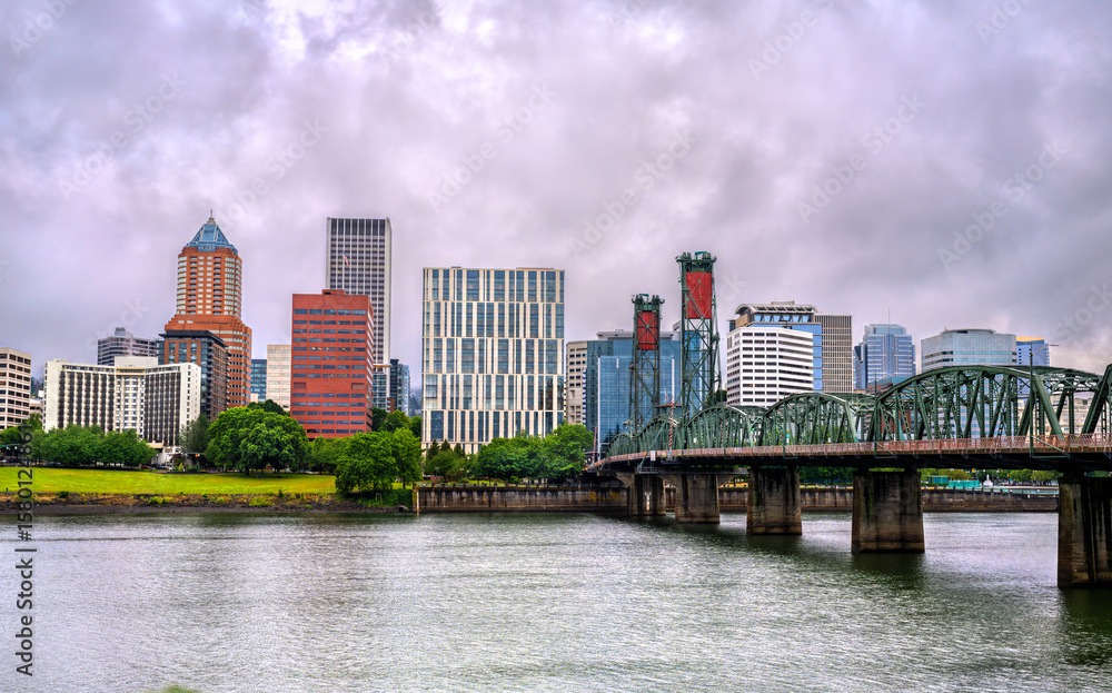 Naklejka premium Panoramic view of Portland's modern downtown skyline under overcast skies in Oregon, USA, with the iconic steel Hawthorne Bridge spanning the Willamette River in the foreground