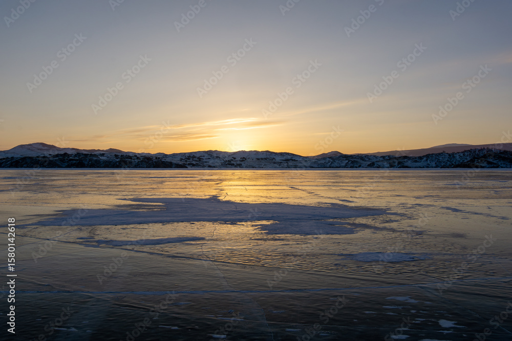 Fototapeta premium Expansive frozen lake reflecting golden sunset hues, with distant snow-capped mountains under a clear sky, creating a serene winter landscape atmosphere