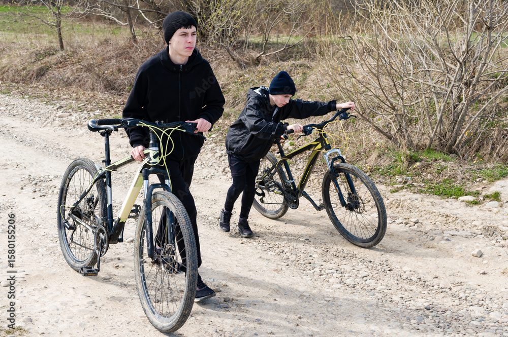 Naklejka premium Two teens walk their bikes down a dirt road, one pushing his bike while the other walks alongside.