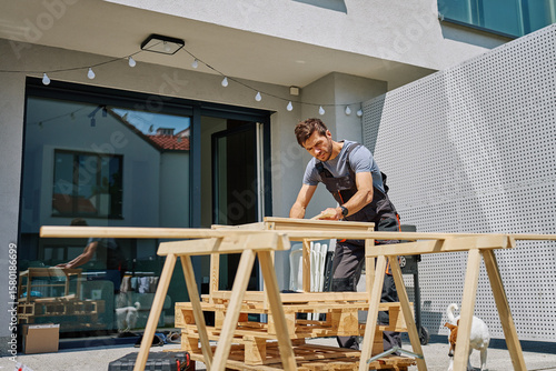 Quadro su tela Man working on outdoor carpentry task, measuring wooden board on sawhorses in ba