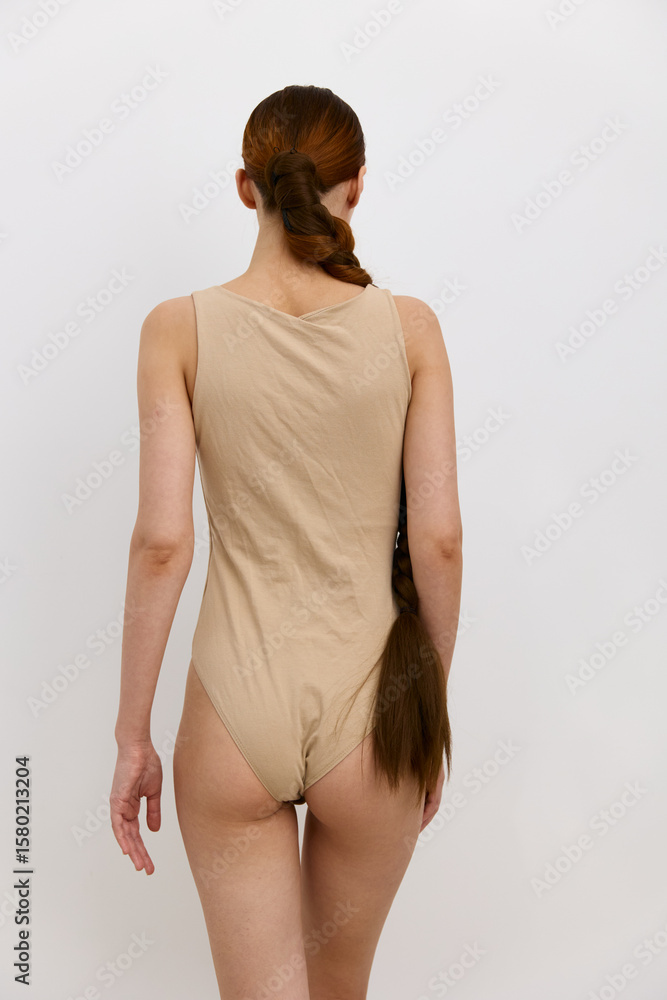Obraz premium woman with long braided hair wearing beige sleeveless bodysuit stands against plain white background, minimalist style and neutral tones emphasize simplicity and elegance.