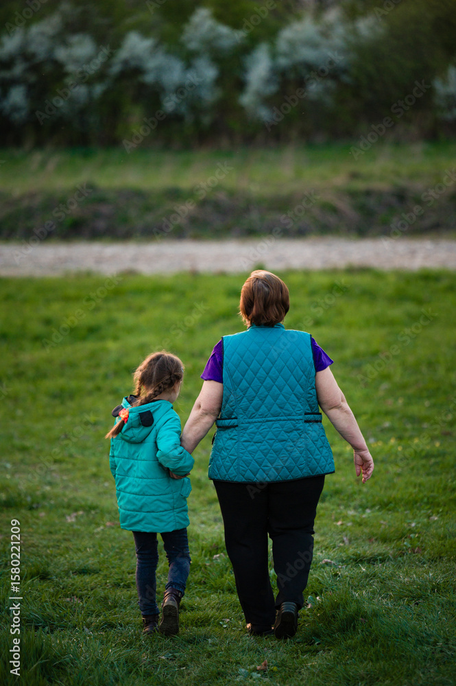 Fototapeta premium Grandmother and granddaughter stroll hand-in-hand across a grassy field, enjoying a peaceful afternoon together.