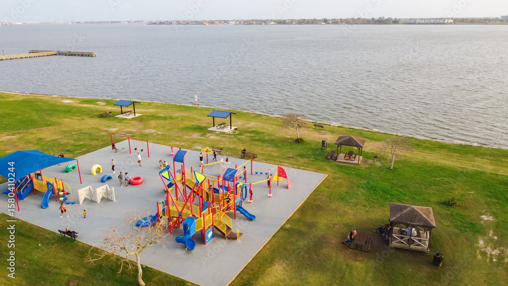 Fototapeta premium Parents and kids enjoy playing at colorful lakefront playground, T-shaped fishing pier stretches into Clear Lake, Seabrook. Scene reflects dynamic lakefront recreation, family-friendly park, Texas