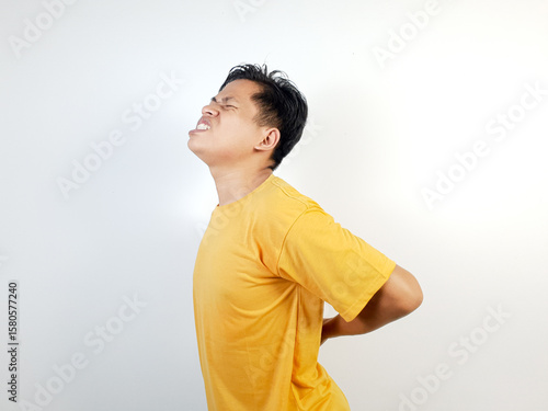 Obraz na płótnie A young Asian man in a yellow shirt struggles with back pain, expressing discomfort against a white background