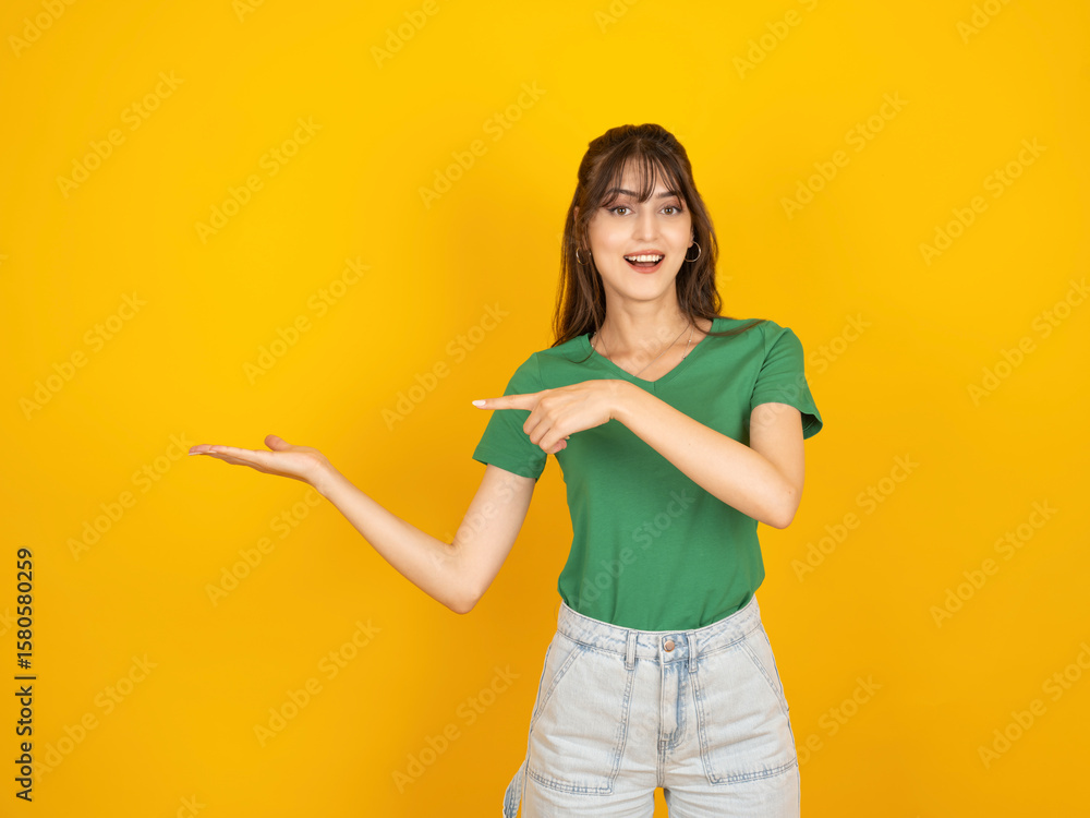 Young excited happy smiling 20s caucasian brunette woman wear green t-shirt and jean standing over isolated yellow background point index finger on empty palm with copy space place mock up. Young excited happy smiling 20s caucasian brunette woman wear green t-shirt and jean standing over isolated yellow background point index finger on empty palm with copy space place mock up.