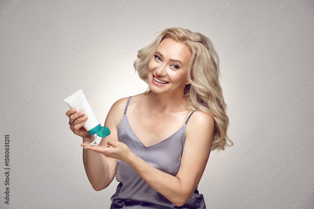 Beauty image of pretty older woman with blonde hair wearing pajamas applying tube of cream for face isolated over grey background. Beauty image of pretty older woman with blonde hair wearing pajamas applying tube of cream for face isolated over grey background.