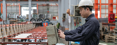 A man in a hardhat and mechanic coverall dust protection suit operates industrial equipment inside a busy factory setting, skillfully managing the machinery.