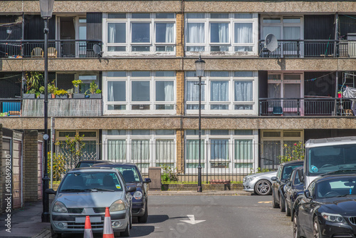 Фотография A council block building with balconies, alongside a street lined with parked ca
