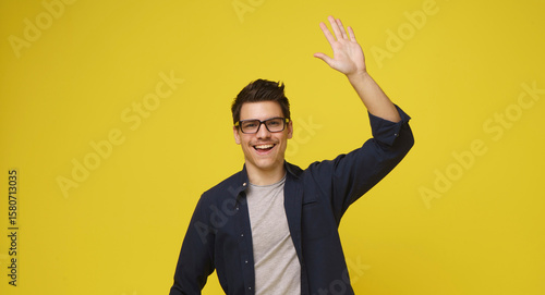 A man wearing glasses smiles and waves with a cheerful expression, standing against a bright yellow background