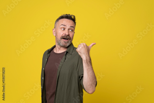 Happy attractive mature 50s hoary senior man isolated over yellow background pointing to the back behind with hand and thumbs up with face excited expression and speaking emotion, smiling confident.