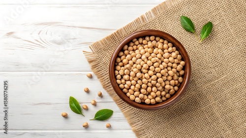 Overhead view of a rustic wooden bowl filled with dried soybeans resting on a burlap cloth with scattered beans and fresh green leaves on a white wooden surface