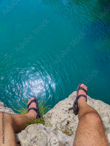 Man in sandals sitting on rocky cliff edge above clear blue sea, legs visible. Ideal for adventure, freedom, travel inspiration, exploration, and nature escape visuals.