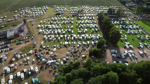 Aerial drone footage flying over a scenic nature campground with multiple RVs, camper vans, and motorhomes parked on a green open space surrounded by trees and natural landscape.

