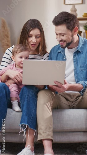 Smiling Caucasian couple sitting close on couch holding baby and looking at laptop. Watching family video or joining video call together. Sharing screen time and enjoying relaxing moment.