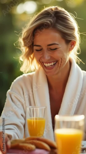 Joyful blonde woman in robe laughing and enjoying breakfast outdoors