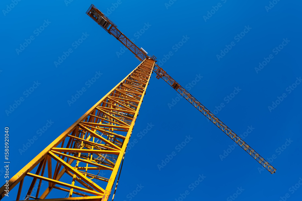 Large orange and yellow construction crane on a building site, cropped from a frog's-eye view in front of a deep blue sky. Steel construction machine for lifting and transporting loads. Large orange and yellow construction crane on a building site, cropped from a frog's-eye view in front of a deep blue sky. Steel construction machine for lifting and transporting loads.