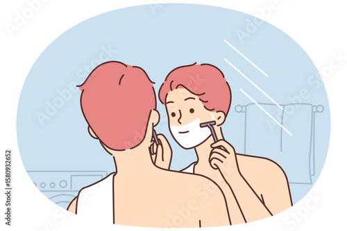 Man shaves face standing in bathroom and looking in mirror doing daily morning hygiene routine