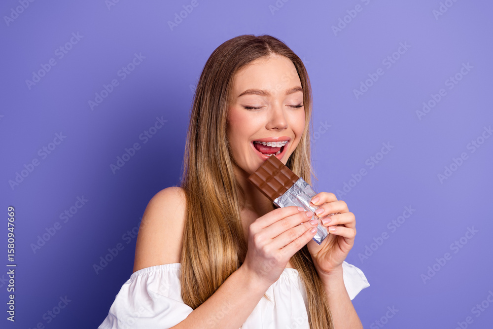 Young woman with braces enjoying a chocolate bar against a vibrant purple background, wearing a trendy casual outfit Young woman with braces enjoying a chocolate bar against a vibrant purple background, wearing a trendy casual outfit