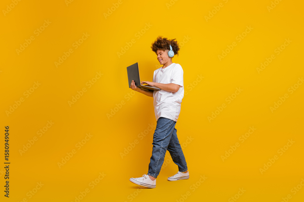Fototapeta premium Confident young boy wearing headphones while holding a laptop, standing against a yellow background and looking cheerful.