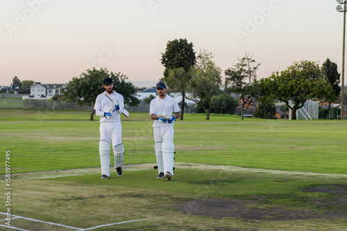 Diverse male teammates walking across cricket pitch wearing batting pads and holding wooden bats