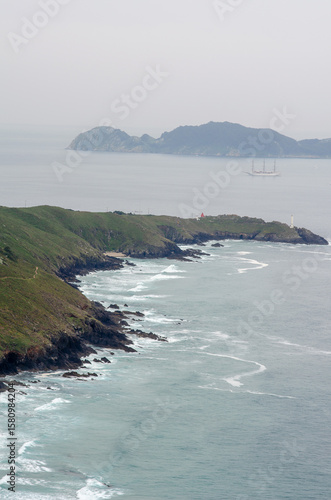 Scenic view of rugged green cliffs meeting the Atlantic Ocean, with a tall ship sailing in the distance under a cloudy sky. Ideal for travel and nature concepts.