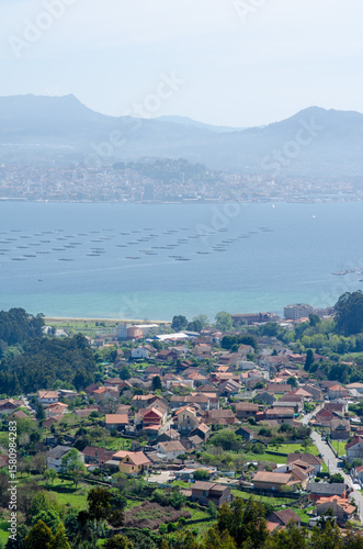 Scenic view of a picturesque Galician village by the sea with distant mountains and mussel farms, capturing peaceful rural life and beautiful spring landscape.
