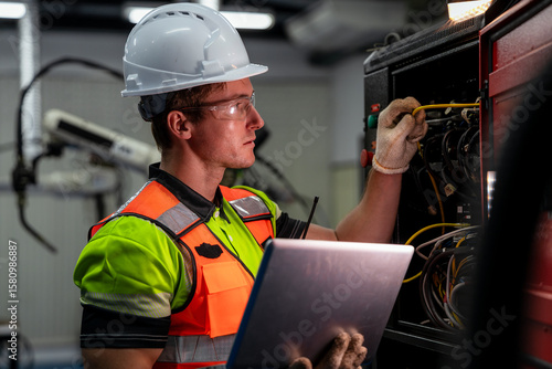 A man in a green and orange vest is working on a laptop. Electrical engineer or technician using a laptop for diagnostics and maintenance of a machine control panel in a factory.