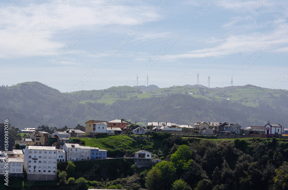 Naklejka premium Peaceful rural village with colorful houses on lush green hills, distant wind turbines, and clear blue sky, capturing sustainable energy and serene countryside life.