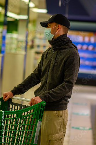 March 09 2022, Cangas - Spain. Adult male shopper wearing a face mask stands with a green cart in a modern grocery store, expressing safety awareness and urban lifestyle.