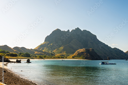 Small boat anchored near the shore of Labuan Bajo, Indonesia, with dramatic hills and a tranquil bay under bright daylight, viewed from a pebbled beach foreground.