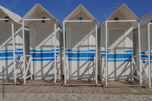 Fototapeta Naklejka Na Ścianę i Meble -  Row of identical white wooden changing cabins standing next to each other on a sandy beach with the sea in the background. Quiet coastal atmosphere, summer vacation, travel.