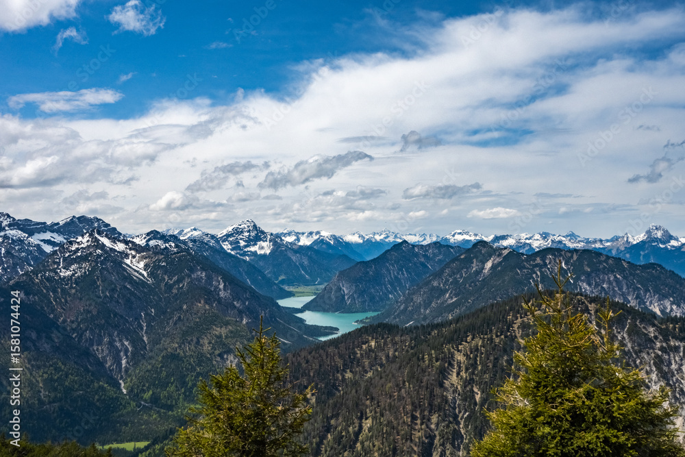Obraz premium Panoramic view of the turquoise Plansee lake nestled in the Alps, seen from the Ammergau mountains. Snow-capped peaks, green forests, and a bright blue sky. Alpine landscape in Tyrol and Bavaria