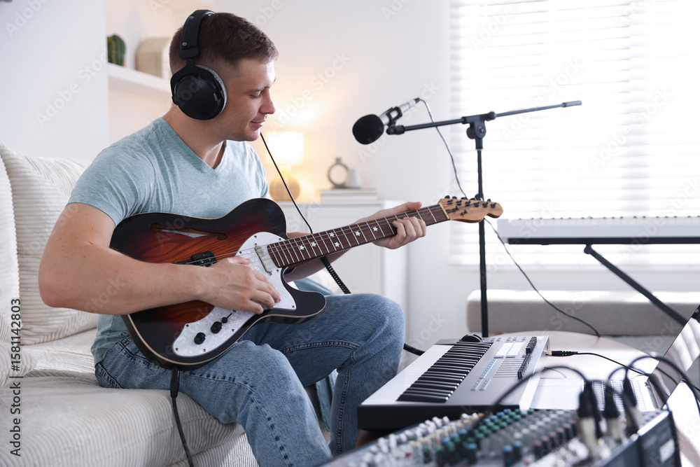 Fototapeta premium Man in headphones working with guitar and MIDI keyboard on sofa in home studio