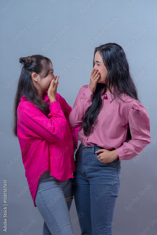 Two young Asian Indonesian women laughing with hands covering their mouths, enjoying a fun moment together while wearing pink shirts and jeans in a grey studio background. Two young Asian Indonesian women laughing with hands covering their mouths, enjoying a fun moment together while wearing pink shirts and jeans in a grey studio background.