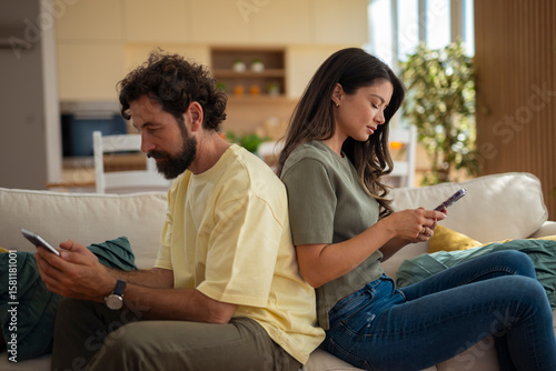 Fényképezés Young couple sitting back to back on a sofa, both absorbed in their smartphones