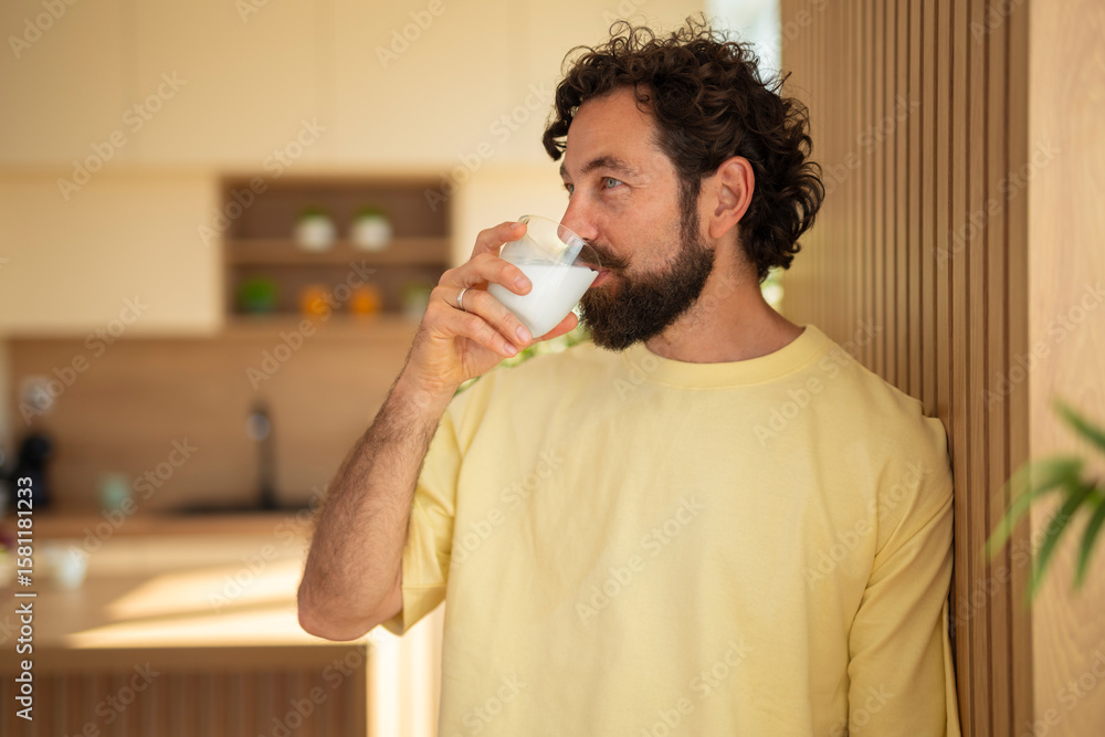 Young man with a beard drinking a glass of milk while leaning against a wooden wall in a modern kitchen. Calm morning atmosphere with soft sunlight and natural interior.
Young man with a beard drinking a glass of milk while leaning against a wooden wall in a modern kitchen. Calm morning atmosphere with soft sunlight and natural interior.